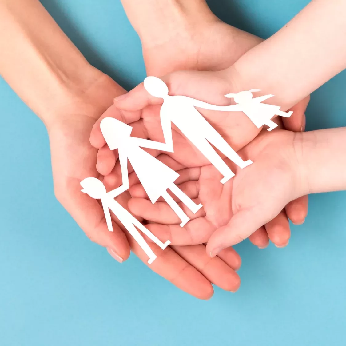 Mains tenant une découpe en papier représentant une famille Symbole de protection et de solidarité familiale : plusieurs mains d’adultes et d’enfants soutiennent une silhouette découpée en papier représentant une famille unie. Une image forte de sécurité, d’amour et de soutien intergénérationnel.