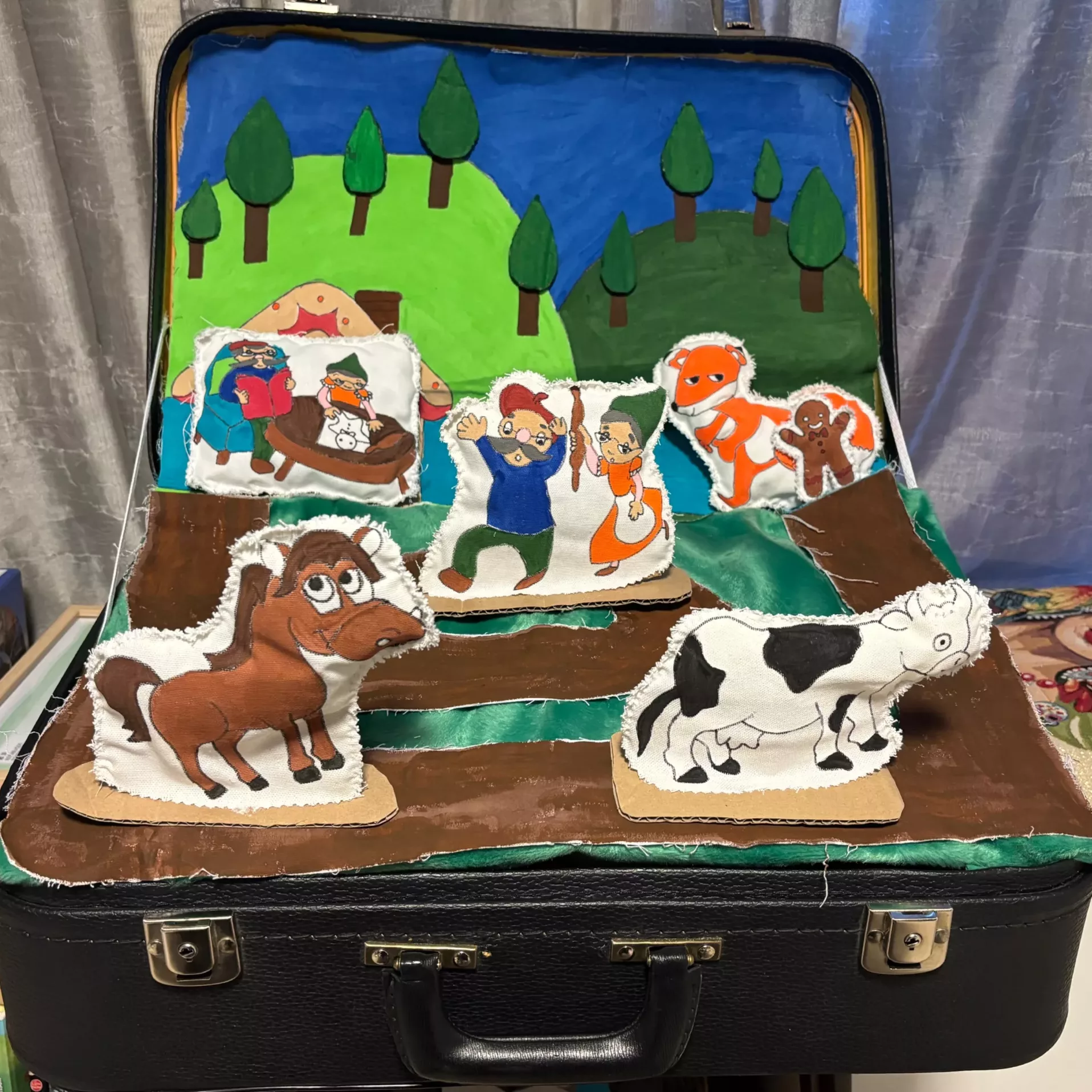 Décor de conte pour enfants avec figurines en tissu représentant des animaux de ferme et des personnages dans une valise Scène ludique installée dans une valise, représentant un décor champêtre fait main avec des figurines en tissu de vaches, chevaux et personnages de contes. Une activité pédagogique et créative pour enfants, idéale pour l'éveil et l'imaginaire.
