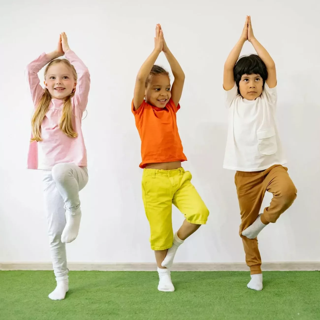 Enfants pratiquant une posture de yoga en équilibre sur un pied Trois enfants souriants effectuent la posture de l’arbre lors d’un cours de yoga, debout sur un tapis vert, illustrant la concentration, l’équilibre et le bien-être à travers une activité physique adaptée aux enfants.